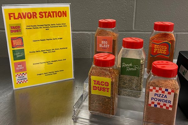 5 bottles of seasoning sit on a stainless steel table with a sign next to it showing what's in them.